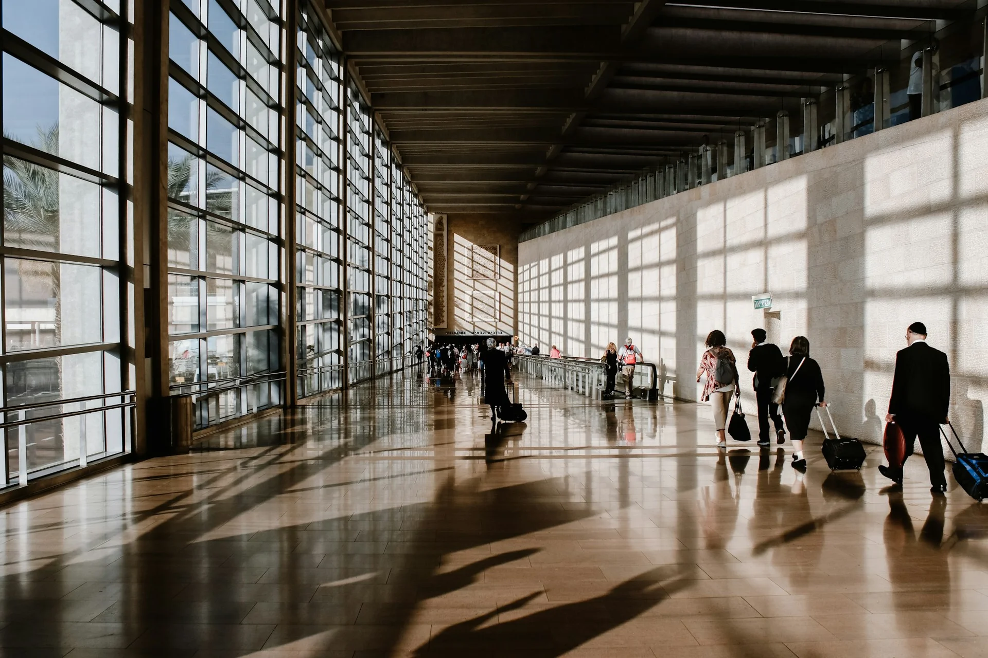 Travelers walking with rolling luggage through a sunlit airport terminal